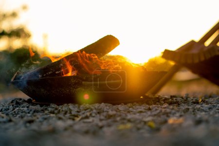 Téléchargez les photos : Gros plan d'un feu de bois au coucher du soleil près d'une confortable maison en bois dans la campagne - en image libre de droit