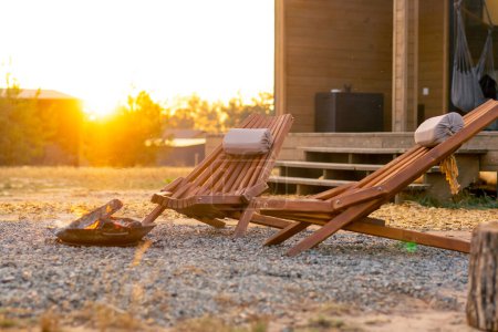 Photo for Outside a fire burns at sunset outside a cosy wooden house with a terrace in a glamping site in steppe - Royalty Free Image