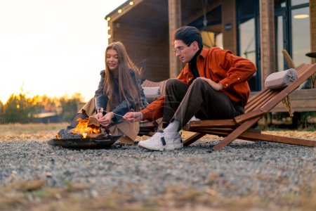 Foto de Joven amante pareja novio y novia sentado junto al fuego cerca de una cabaña de madera en el campo y tostado malvaviscos - Imagen libre de derechos