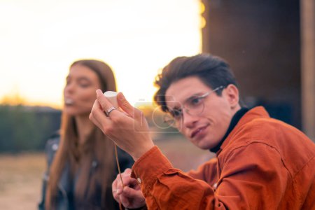 Photo for Close-up shot of a young stylish guy with his girlfriend tasting marshmallow cooked on campfire - Royalty Free Image
