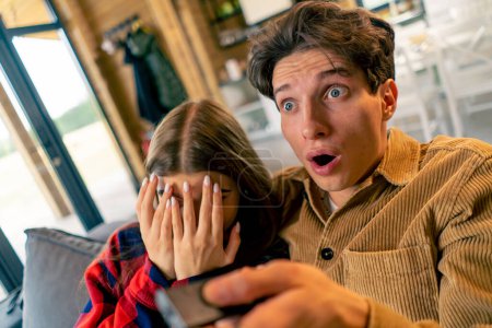 Téléchargez les photos : Un jeune couple en regardant un film d'horreur Le mec est surpris et la fille se couvre le visage avec ses mains dans la peur - en image libre de droit