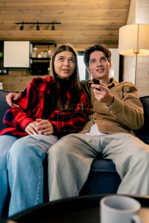 Foto de Sonriente pareja enamorada de un chico y una chica están sentados juntos en el sofá y viendo una película de comedia en una acogedora casa de madera - Imagen libre de derechos