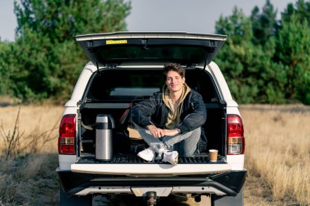 Téléchargez les photos : Un type cher est assis dans le coffre ouvert de sa voiture avec un thermos et un verre de thé à l'extérieur dans un champ - en image libre de droit