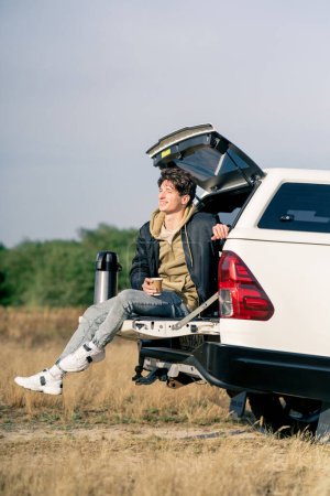 Foto de Un tipo pensativo se sienta en el maletero abierto de su coche con un termo y un vaso de té al aire libre en un campo - Imagen libre de derechos