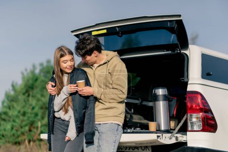 Foto de Un tipo le pone una chaqueta a su novia con una taza de té mientras está de pie cerca del maletero abierto del coche en la naturaleza. - Imagen libre de derechos