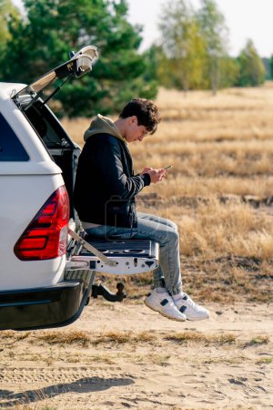 Photo for A young guy sits in the open trunk of a car with phone and studies the area during a stop in a meadow - Royalty Free Image