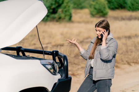 Foto de El coche se averió en la carretera en el bosque y una joven llama por teléfono pidiendo ayuda para arreglarlo. - Imagen libre de derechos