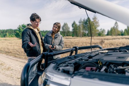 Photo for A guy driver helps a girl fix her car in a field and forest during a trip by opening the hood - Royalty Free Image