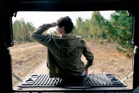Foto de La parte posterior de un tipo que se sienta en el maletero de un coche y admira la vista del prado de campo y el bosque a lo largo de la carretera - Imagen libre de derechos