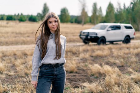 Téléchargez les photos : Une belle fille se tient dans un champ sur fond de voiture avant un voyage à travers la forêt - en image libre de droit