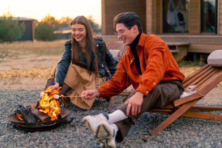 Foto de Joven amante pareja novio y novia sentado junto al fuego cerca de una cabaña de madera en un campo y tostado malvaviscos - Imagen libre de derechos