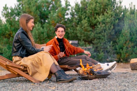 Téléchargez les photos : Un couple d'amis gars et fille assis ensemble autour d'un feu de camp dans un bois faire des guimauves et bavarder animément - en image libre de droit