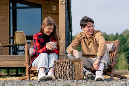 Foto de En la terraza cerca de una gran casa pareja enamorada se sienta con tazas de té en sillas de madera y se comunica entre sí - Imagen libre de derechos
