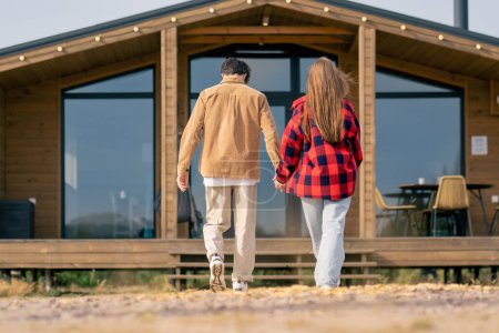 Photo for A guy and a girl walk together holding hands into wooden house with a large panoramic windows - Royalty Free Image