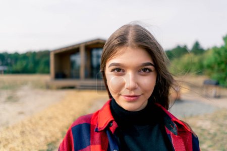 Foto de Retrato de cerca de una hermosa chica mirando directamente a la cámara y sonriendo sobre el fondo de la casa de madera en el bosque - Imagen libre de derechos