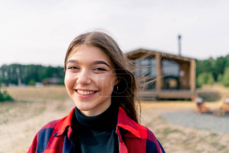 Foto de Retrato de cerca de una hermosa chica mirando directamente a la cámara y sonriendo sobre el fondo de la casa de madera en el bosque - Imagen libre de derechos