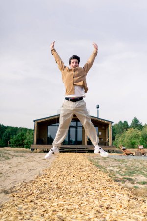 Téléchargez les photos : Un jeune homme joyeux rebondissant de joie devant un grand chalet en bois dans les bois - en image libre de droit