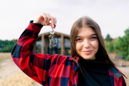 Photo for A girl holds a bunch of keys to her new home against the backdrop of a wooden cottage in the woods in the countryside - Royalty Free Image
