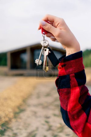 Téléchargez les photos : Gros plan de la main d'une femme tenant les clés de sa maison de campagne en bois récemment achetée dans un bois - en image libre de droit