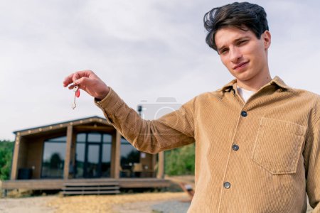 Photo for A guy holds a bunch of keys to his new house in the background of a wooden cabin in a woods in the countryside - Royalty Free Image