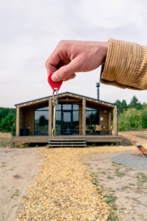 Téléchargez les photos : Gros plan de la main d'un homme tenant les clés de sa maison en bois de banlieue nouvellement achetée dans un bois - en image libre de droit