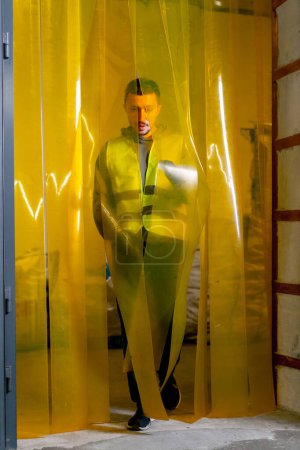 Foto de Un empleado de la estación de reciclaje de residuos con un chaleco de trabajo camina a través de una cortina de silicona en la puerta de la planta - Imagen libre de derechos