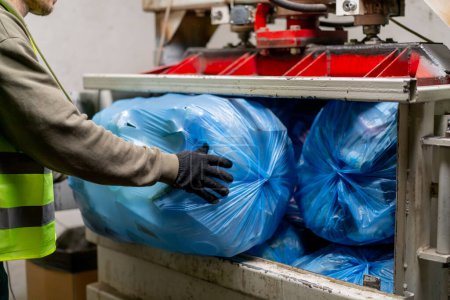 Foto de Primer plano de las manos masculinas en guantes cargando grandes bolsas de basura en una máquina compactadora de basura especial para la eliminación de residuos - Imagen libre de derechos
