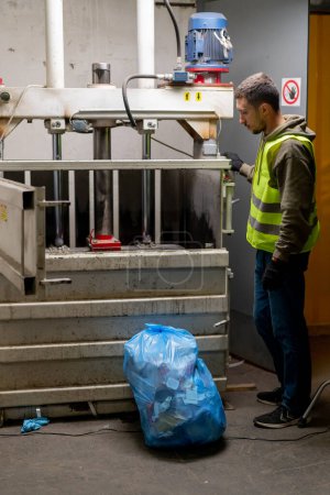 Foto de Un empleado de la planta de reciclaje en uniforme pone bolsas de basura en una máquina de prensa especial para la eliminación de residuos - Imagen libre de derechos