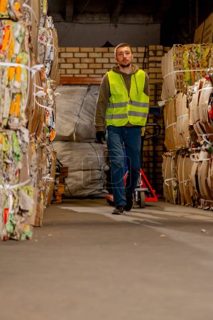 Téléchargez les photos : Un travailleur vêtu d'un gilet de sécurité et de gants marche dans un couloir avec un transpalette hydraulique pour transporter de grandes boîtes d'articles à recycler - en image libre de droit