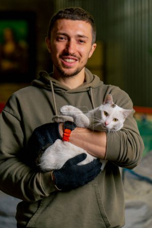 Foto de Retrato de un trabajador en una estación de reciclaje de residuos sosteniendo a un hermoso gato local en sus brazos y sonriendo - Imagen libre de derechos