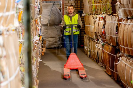 Photo for A worker in a safety vest and gloves walks down a hallway with a hydraulic pallet truck for transporting large boxes of items for recycling - Royalty Free Image