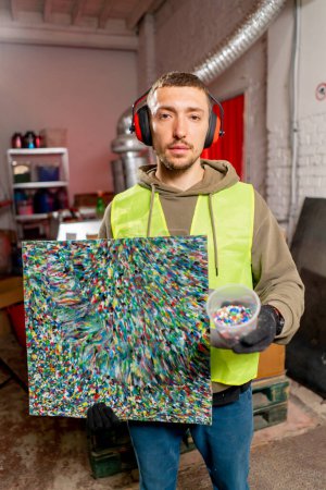 Photo for A factory employee holds a painting made from shredded plastic bottle caps at a waste recycling station - Royalty Free Image