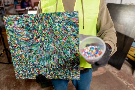 Foto de Un primer plano de una pintura hecha de tapas de plástico reciclado triturado colocadas en un vaso junto a ella - Imagen libre de derechos