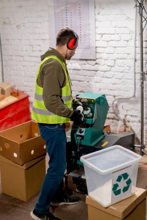 Téléchargez les photos : Un employé d'une usine de recyclage des déchets en uniforme et un casque verse des bouchons en plastique déchiqueté dans un conteneur pour recyclage - en image libre de droit