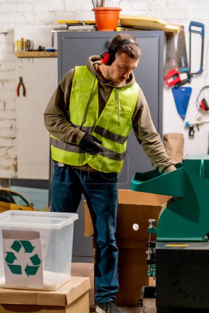 Foto de Un empleado de la planta de reciclaje de residuos en uniforme y auriculares vierte tapas de plástico trituradas en un contenedor para reciclar - Imagen libre de derechos
