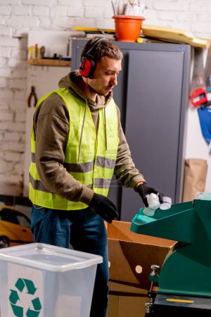 Foto de Un empleado de la estación de reciclaje de residuos que usa auriculares que cancelan el ruido enciende una máquina especial para triturar tapas de plástico usadas - Imagen libre de derechos