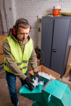 Téléchargez les photos : Un employé d'une station de recyclage des déchets portant un casque anti-bruit allume une machine spéciale pour déchiqueter les bouchons en plastique usagés - en image libre de droit