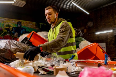 Foto de Un empleado con uniforme y guantes en una estación de reciclaje de residuos clasifica y clasifica la basura en una línea de clasificación - Imagen libre de derechos