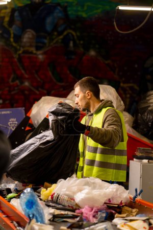 Foto de Clasificador masculino enfocado con un chaleco protector y guantes descarga residuos de una bolsa de basura en una línea de clasificación de basura - Imagen libre de derechos
