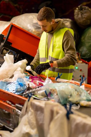 Photo for Side view of a sorter in gloves and a protective vest who sorts garbage on a special sorting line while working at a waste disposal station - Royalty Free Image