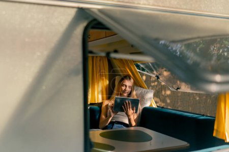 Foto de Mientras viaja en un remolque una joven se acuesta en el sofá junto a la ventana del coche y lee un libro en una tableta - Imagen libre de derechos