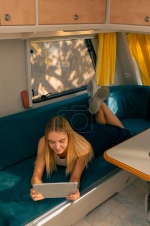 Photo for A girl lies on her stomach on a sofa in a camper smiles and watches a movie on a tablet while traveling - Royalty Free Image