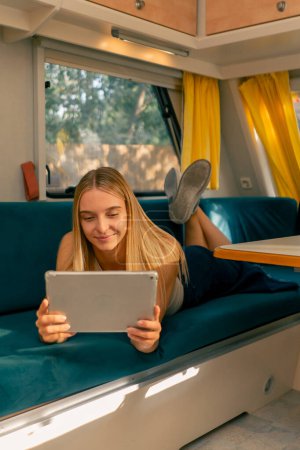 Photo for A girl lies on her stomach on a sofa in a camper smiles and watches a movie on a tablet while traveling - Royalty Free Image