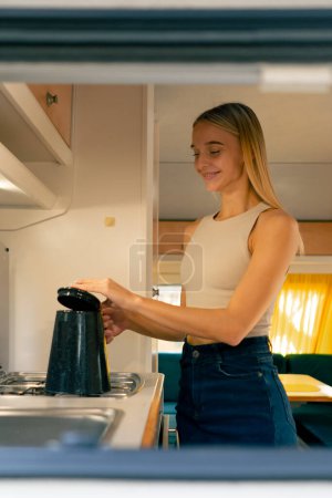 Téléchargez les photos : Une fille met une bouilloire sur la cuisinière pour chauffer l'eau et préparer le thé le matin dans la cuisine d'un mobil-home - en image libre de droit