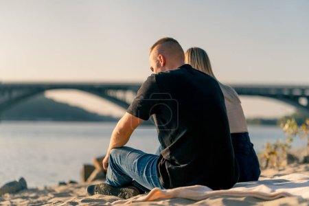Foto de Silueta de una pareja enamorada sentada en la orilla arenosa del río admirando el sol poniente y el hermoso paisaje de la naturaleza - Imagen libre de derechos
