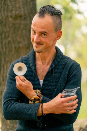 Téléchargez les photos : Maître de thé souriant regardant la caméra et montrant beau bol de thé en céramique sur fond d'arbres au camping de campagne - en image libre de droit