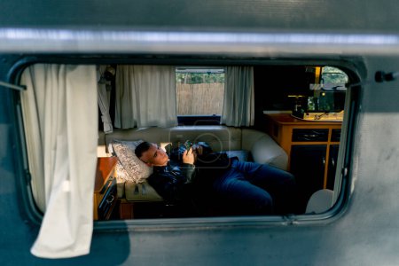 Foto de Un hombre triste cansado yace deprimido en el sofá junto a la ventana del remolque y mira indiferentemente la pantalla de su teléfono - Imagen libre de derechos