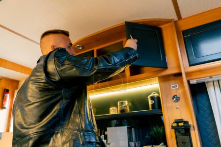 Téléchargez les photos : Un homme ouvre tiroirs et armoires dans la cuisine à la recherche d'une tasse pour faire du thé dans un camping-car - en image libre de droit