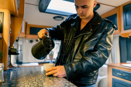 Photo for A man in the kitchen of a motorhome pours boiling water from a kettle into a cup to make morning tea - Royalty Free Image