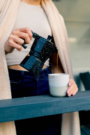 Téléchargez les photos : Gros plan de la main d'une femme tenant un verre et versant du café fraîchement infusé d'une cafetière sur une terrasse avec des fleurs - en image libre de droit